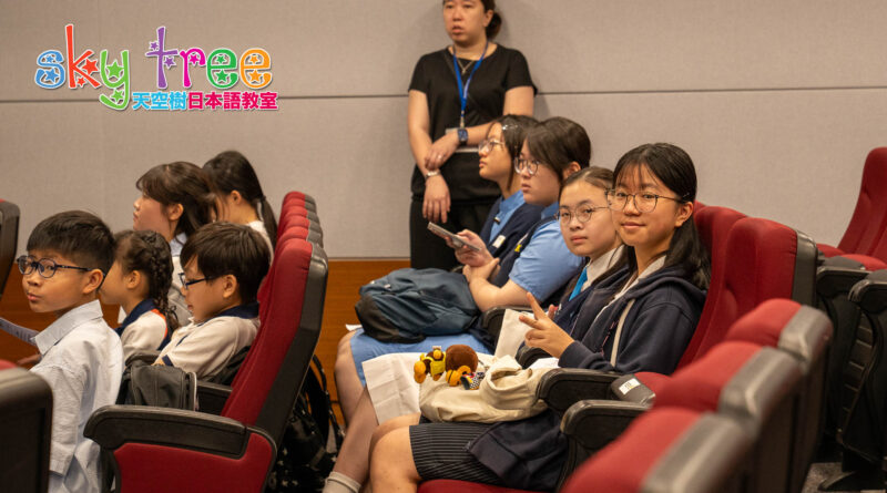 DSC05051 | 天空樹日本語教室 Group of students sitting in red theater seats, looking toward the front; a teacher stands at the back against a beige wall with a colorful Sky Tree logo in the corner.
