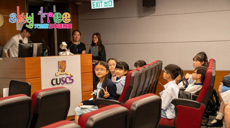 DSC05057 | 天空樹日本語教室 Students seated in a lecture hall, facing the front where presenters stand beside a desk with a CSUCS sign and equipment, some looking at the camera and smiling.