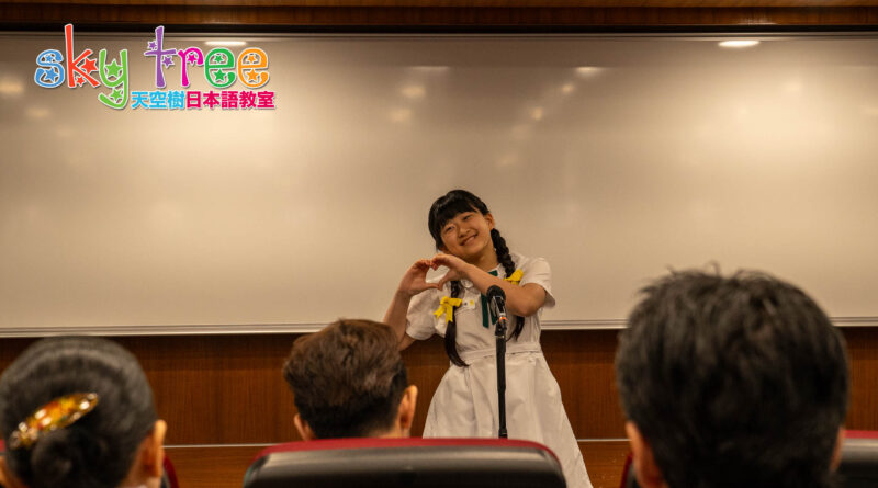 DSC05070 | 天空樹日本語教室 Girl in a white dress with yellow ribbons forms a heart shape with her hands at a microphone on a stage, audience in the foreground.