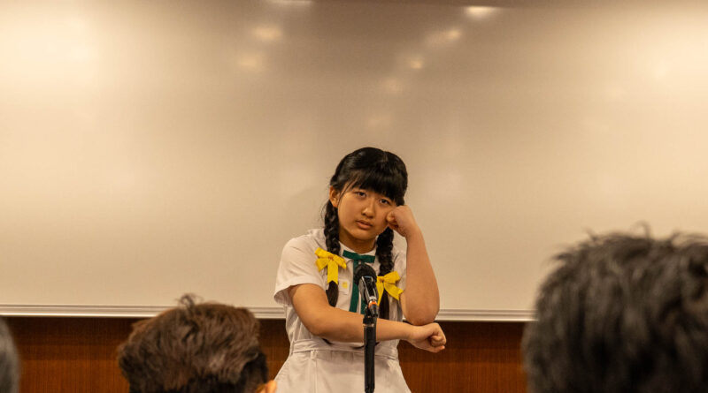 DSC05072 | 天空樹日本語教室 Girl with braided hair standing at a lectern in a classroom, speaking into a microphone, with a colorful 'Sky Tree' logo and Japanese text on the wall behind her.
