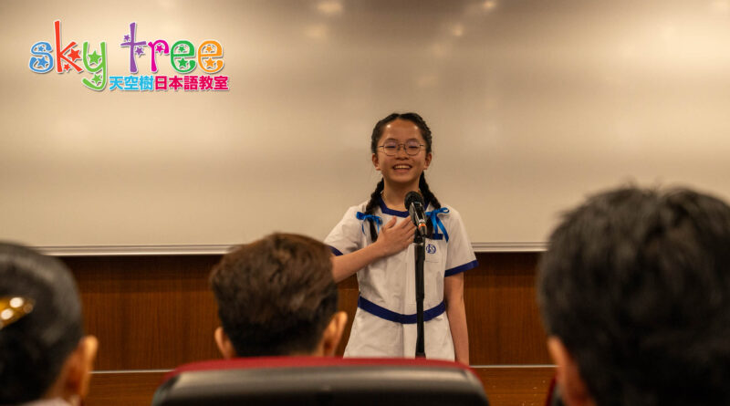 DSC05077 | 天空樹日本語教室 Young girl in a white and blue school uniform speaks into a microphone on stage, audience members visible in the foreground, with a colorful 'Sky Tree' logo in the upper left.