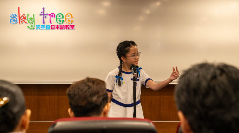 DSC05083 | 天空樹日本語教室 Young girl in a white and blue uniform speaks into a microphone at a podium, with a colorful logo in the top-left corner and seated audience in front.