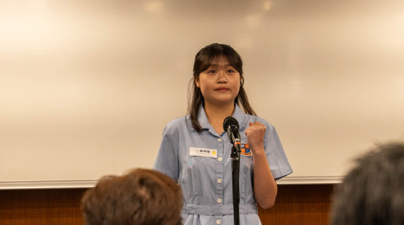 DSC05095 | 天空樹日本語教室 Woman in a light blue uniform speaks into a microphone at a podium, with audience members visible in the foreground in a classroom setting.