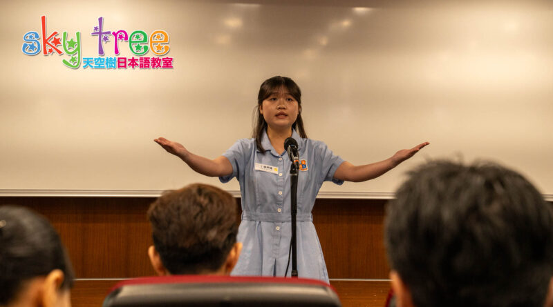 DSC05099 | 天空樹日本語教室 Young woman in a light blue dress speaks at a microphone with arms outstretched in a classroom, audience visible in the foreground.