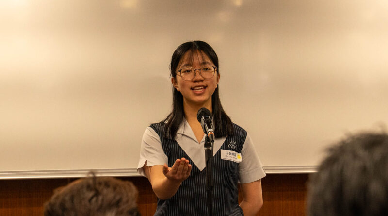 DSC05119 | 天空樹日本語教室 Female presenter in a pinstripe dress speaks into a microphone at the front of a lecture hall, audience blurred in the foreground, logo in the top left corner.