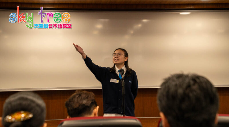DSC05143 | 天空樹日本語教室 Female student giving a presentation at a classroom, gesturing with her right hand, microphone in front, audience visible in foreground; colorful 'Sky Tree' logo on the wall behind her.