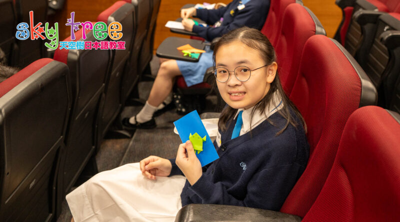 DSC05151 | 天空樹日本語教室 Girl in a navy school uniform with glasses sits in red seats, holding a blue card with a green origami frog.