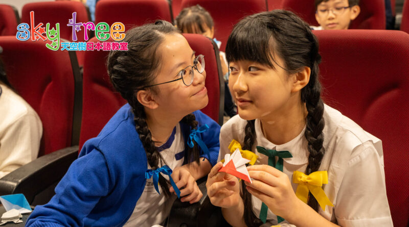 DSC05155 | 天空樹日本語教室 Two schoolgirls with braided hair and colorful bows share a smile while making origami in red theater seats.