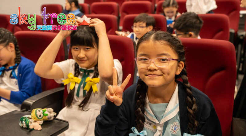 DSC05157 | 天空樹日本語教室 Two smiling girls sit in red theater seats; the girl on the right wears glasses and braids, making a peace sign, while the girl on the left adjusts a paper crown. A colorful 'Sky Tree' logo is in the top-left corner.