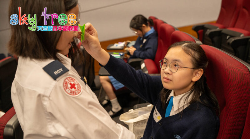 DSC05161 | 天空樹日本語教室 Volunteer in a white uniform places a green bandage on a young woman's forehead during a first-aid training in a red-seat auditorium.
