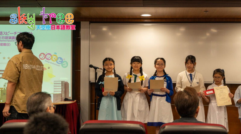 DSC05166 | 天空樹日本語教室 Five girls in school uniforms stand on a stage holding certificates during a ceremony, with an audience in front.