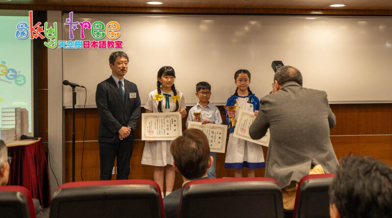 DSC05196 | 天空樹日本語教室 Three students in school uniforms stand on a stage, holding certificates and trophies while a presenter and photographer look on at an award ceremony.