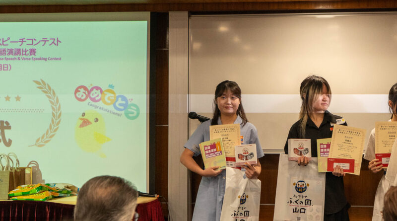 DSC05228 | 天空樹日本語教室 Three young women stand on a stage holding certificates and white bags, with a projection screen displaying Japanese text to the left and an audience in front.