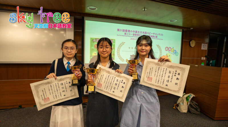 DSC05286 | 天空樹日本語教室 Three young girls in school uniforms proudly hold large certificates and gold trophies during an awards ceremony, with a projector screen and colorful logo in the background.