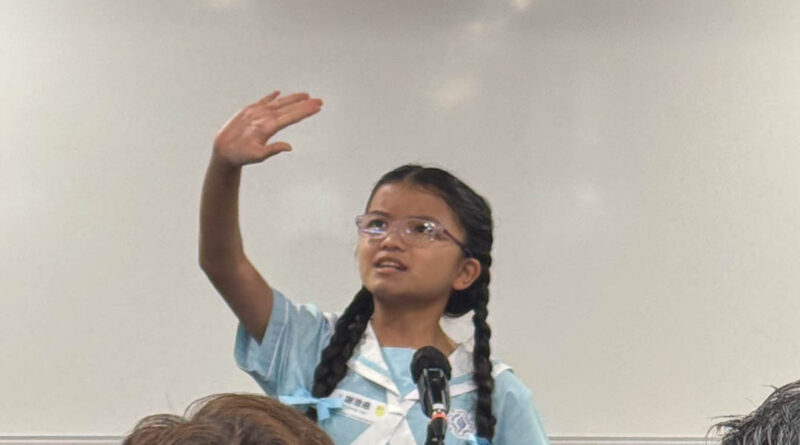 IMG_2430 | 天空樹日本語教室 Young girl in a blue school uniform speaks into a microphone, raising her hand to answer.