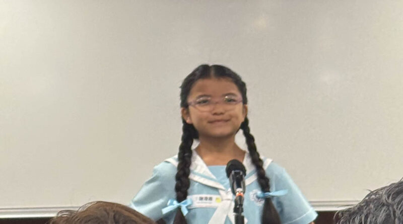 IMG_2431 | 天空樹日本語教室 Young girl in a light blue school uniform speaks into a microphone during a classroom presentation, with audience members in the foreground and a colorful logo in the top corner.